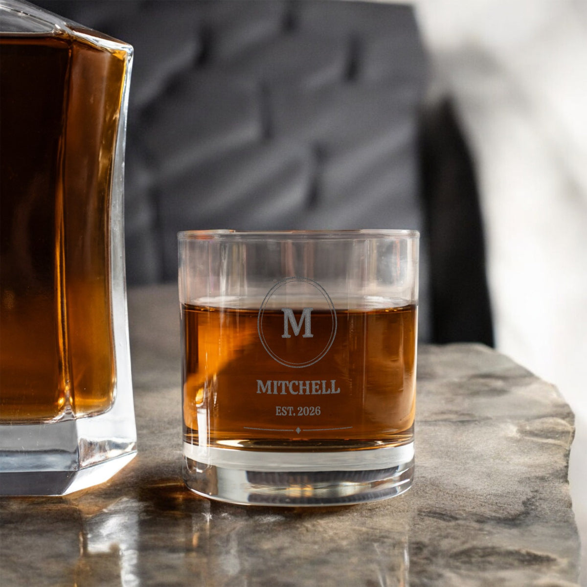 Etched monogram whiskey glass beside a decanter on a stone surface