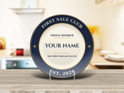 Navy and gold First Sale Club keepsake plate with a personalized name at the center, on a light wood counter